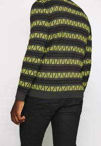 Pull-over en maille gris foncé avec des motifs géométriques jaunes. Doté de poignets et d'un ourlet côtelés ; le tissu semble doux avec une finition texturée.