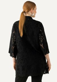 Kimono negro de encaje con intrincados patrones florales y bordes festoneados. Cuenta con mangas de ¾ y un tejido ligero y texturizado.