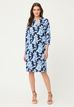 Kobieta stojąca w granatowej sukience do kolan z dużymi niebiesko-białymi kwiatowymi wzorami, ubrana w brązowe sandały na koturnie z paskami.