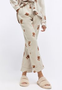 Lichtbeige pyjamabroek met een herhaald teddybeerprint, voorzien van een elastische tailleband en een licht uitlopende snit, gedragen met fluffy slippers.