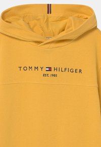 Żółta bluza z kapturem z teksturowanego materiału. Na froncie nadrukowane logo "TOMMY HILFIGER" oraz "EST. 1985" w granatowym kolorze.