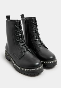 Botas de tobillo de cuero negro con un diseño de cordones, cremallera lateral y costuras blancas contrastantes en la gruesa suela negra. Superficie texturizada.
