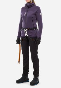 Femme portant une veste violette, un harnais d'escalade noir et des gants, tenant un piolet orange, prête pour l'alpinisme ou l'escalade sur glace.