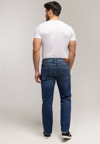 Mężczyzna w białej koszulce, niebieskich dżinsach z tylnymi kieszeniami i czarnych trampkach, stojący tyłem do kamery.