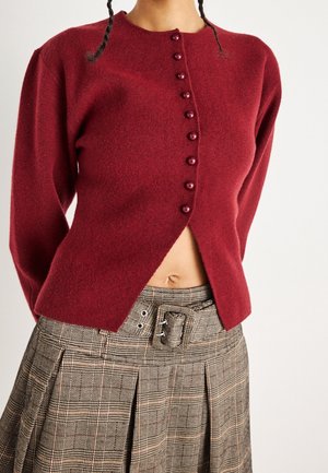 Le modèle porte un cardigan bordeaux boutonné sur le devant et une jupe plissée marron à carreaux avec une ceinture à boucle.