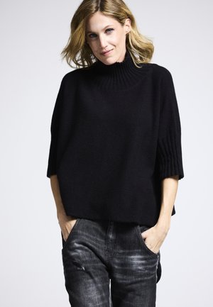 Schwarzer gestrickter Pullover mit hohem Kragen, kurzen Ärmeln und lockerer Passform, kombiniert mit grauen Jeans, die eine dezente Textur und Taschen aufweisen.