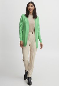 Cardigan vert, top beige et pantalon beige. Le cardigan a une texture côtelée, un devant ouvert et des manches longues. Des bottines noires à la cheville complètent la tenue.