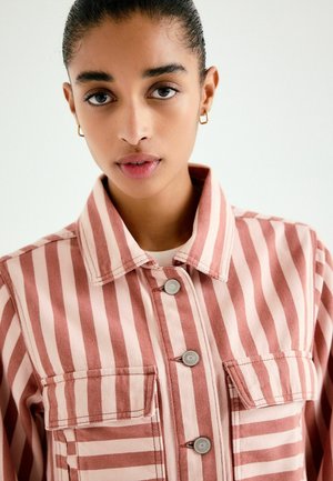 Woman with smooth dark hair in a high bun wears small gold hoop earrings and a pink and white vertically striped button-up jacket.