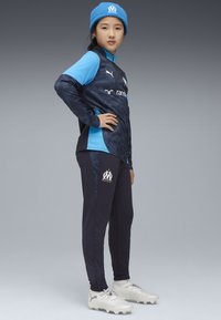 Completo sportivo blu e navy con una maglietta a maniche lunghe testurizzata e pantaloni tapered. Presenta un logo e dettagli bianchi. Scarpini da calcio argentati e berretto di maglia.