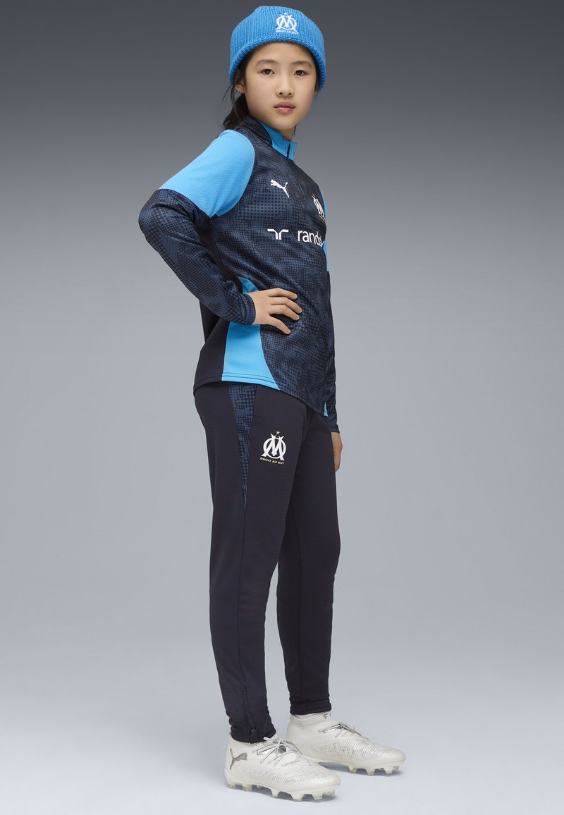 Completo sportivo blu e navy con una maglietta a maniche lunghe testurizzata e pantaloni tapered. Presenta un logo e dettagli bianchi. Scarpini da calcio argentati e berretto di maglia.