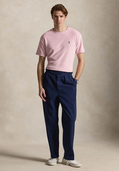 Νέος άνδρας στέκεται με τα χέρια στις τσέπες των ναυτικών παντελονιών, φορά ροζ πουκάμισο με κοντά μανίκια, λευκά αθλητικά παπούτσια και ρολόι χειρός, μπροστά σε μονόχρωμο φόντο.