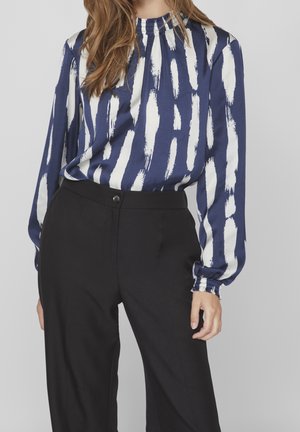 Femme portant un chemisier bleu marine à manches longues avec des rayures blanches en coups de pinceau et un pantalon noir taille haute, debout devant un fond uni.