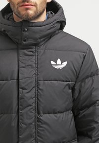 Svart vadderad jacka med hög krage, tryckknappar och en vit logotyp på vänster bröst. Materialet ser slätt ut med en mjuk textur.