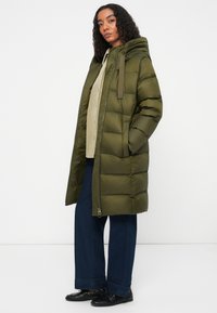 Abrigo largo acolchado verde oliva con capucha y cordón, con cierre delantero y bolsillos laterales, combinado con vaqueros de pierna ancha azules y zapatos negros.