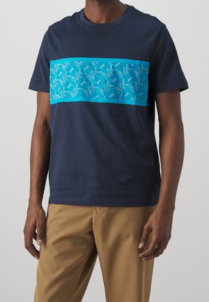 T-shirt en coton bleu marine avec un panneau horizontal cyan orné d'un motif géométrique. Manches courtes et col rond. Porté avec un pantalon beige.