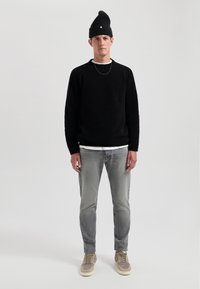Schwarzer Strickpullover, graue Jeans und beigen Sneakers. Die Accessoires bestehen aus einer schwarzen Mütze mit grünem Akzent und einer schlichten goldenen Kette.