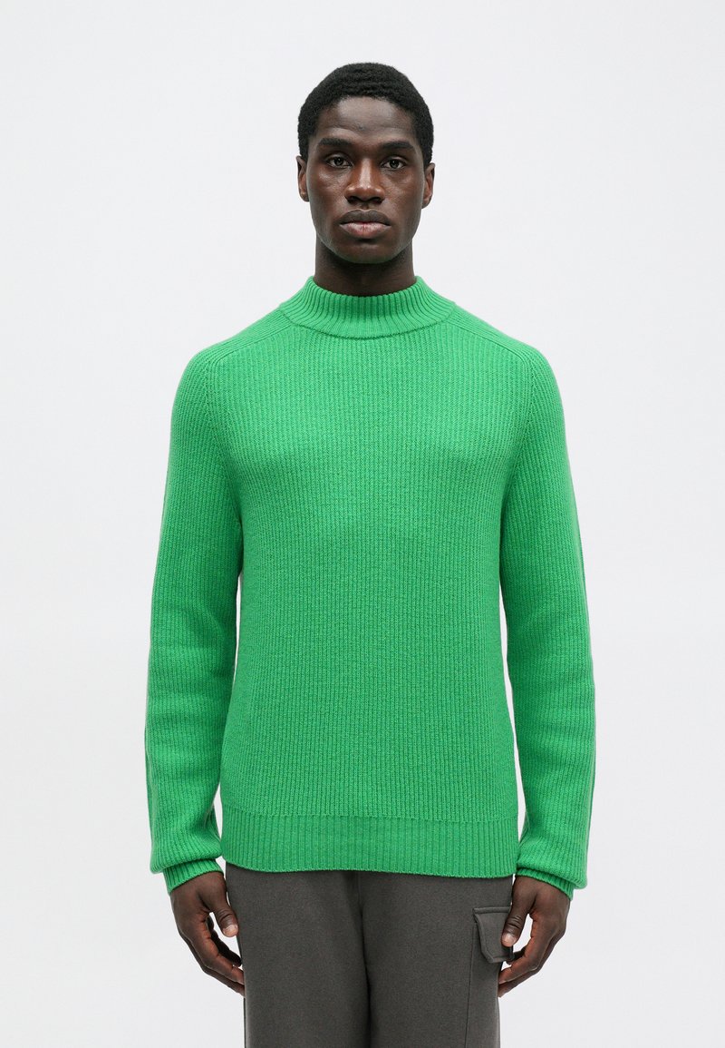 Homme portant un pull côtelé vert vif à manches longues et un pantalon gris avec une poche latérale, sur un fond blanc uni.