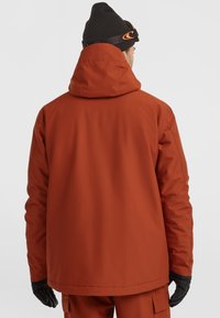 Oranje waterdichte jas met capuchon, textuur oppervlak en elastische manchetten. Gekozen in combinatie met een donkere beanie en zwarte handschoenen. Eenvoudig, functioneel ontwerp.