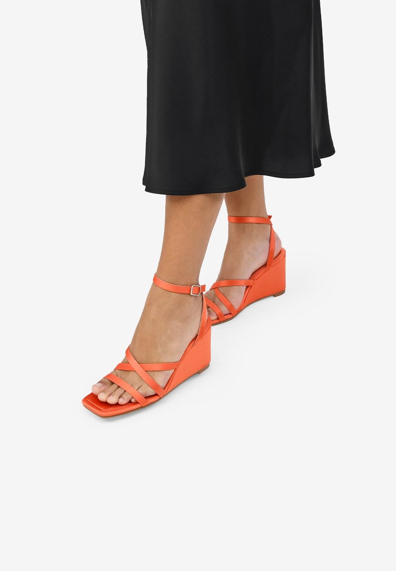 Sandalias de cuña naranja con múltiples correas entrelazadas, diseño de punta abierta y hebillas en el tobillo. Acabado mate con suela plana.