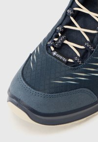 Tumši zila sporta apavi ar teksturētu audumu, gaiši pelēkām detaļām, krēmkrāsas šņorēm un redzamu GORE-TEX etiķeti ūdensnecaurlaidībai.