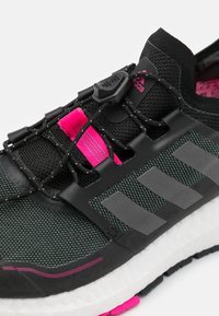 Svart träningssko med en texturerad ovandel, rosa detaljer, elastiska snören och en vadderad vit sula med svart mönster.