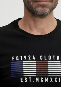Fekete póló grafikus dizájnnal, kék, fehér és piros csíkokkal, valamint a "FQ1924 CLOTHING EST. MCMXXI" felirattal a mellkason.