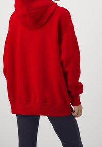 Une personne portant un sweat à capuche rouge surdimensionné et un pantalon foncé ajusté, vue de dos contre un fond clair uni.