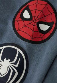 T-krekls ar diviem zirnekļu logo dizainiem: sarkans Spider-Man emblēma ar tīkla rakstu un melna zirnekļa ikona uz pelēka fona.