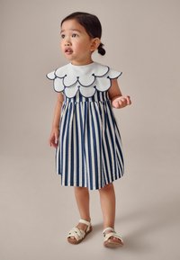 Gestreiftes Kleid mit einem gewellten weißen Kragen, der in Marineblau umrandet ist. Der Stoff zeigt senkrechte Streifen in Blau und Creme. Kombiniert mit hellen Sandalen.