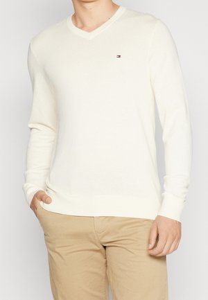 Man draagt een crèmekleurige V-hals trui met een klein logo op de borst en beige broek, hand in de zak, tegen een effen lichte achtergrond.