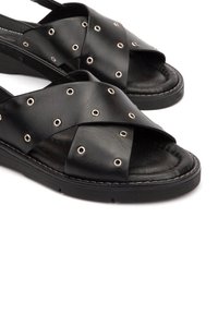 Las sandalias de cuero negro cuentan con tiras entrelazadas con ojales metálicos. La suela tiene una textura que proporciona agarre y comodidad, con un diseño de perfil bajo.