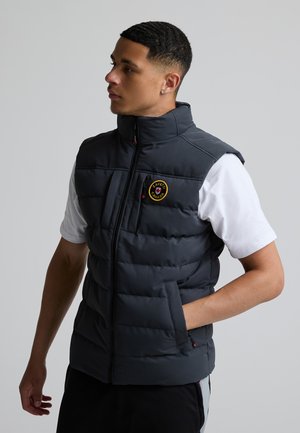 Ung mand iført en sort polstret vest over en hvid t-shirt, stående med den ene hånd i lommen og kigger til siden mod en ensfarvet baggrund.