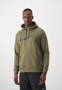 Sudadera de Nike color verde oliva con un gran logo de swoosh negro, bolsillo tipo canguro, capucha con cordón y puños de canalé, combinada con pantalones negros.