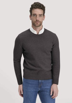 Dunkelgrauer Strickpullover mit Rundhalsausschnitt und gerippten Bündchen, über einem weißen Kragenshirt getragen. Kombiniert mit blauen Jeans, einfarbiger Hintergrund.