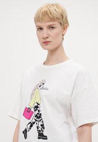 Hvit t-skjorte med et grafisk motiv av en kvinne med langt hår, iført en gul topp og svarte blomstrete bukser, og bærende på en rosa veske. Korte ermer.