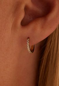 Boucles d'oreilles en or en forme de créole, ornées d'une rangée de petits strass clairs le long du bord avant, mises en valeur par un gros plan d'une oreille humaine.