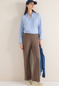 Femme portant un chemisier bleu à manches longues, un pantalon large marron, des baskets blanches, une casquette marine, tenant une veste en denim bleu, debout contre un mur beige.