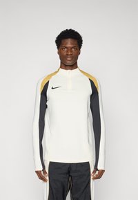 Dugih rukava sportska majica u krem boji, s crnim i zlatnim detaljima. Sadrži ovratnik na pola zatvarača i Nike logo na prsima.
