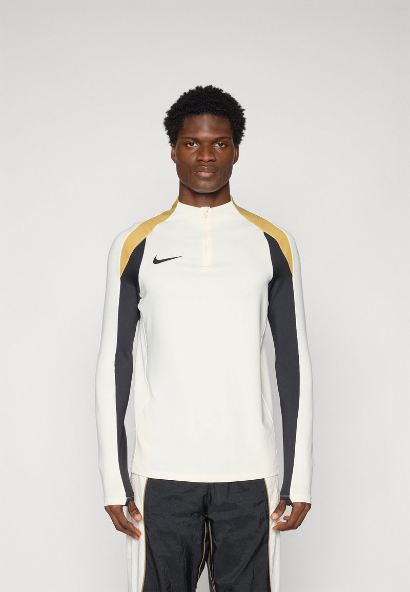 Dugih rukava sportska majica u krem boji, s crnim i zlatnim detaljima. Sadrži ovratnik na pola zatvarača i Nike logo na prsima.