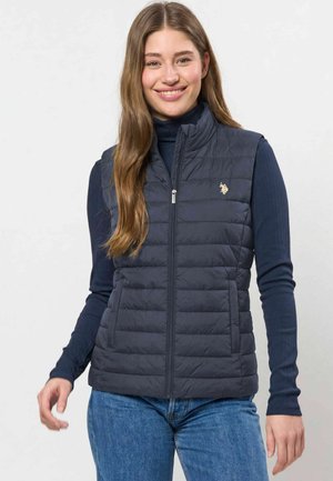 Jeune femme souriante, portant un gilet sans manches matelassé bleu marine sur un haut sombre à manches longues et un jean bleu, debout devant un fond uni.