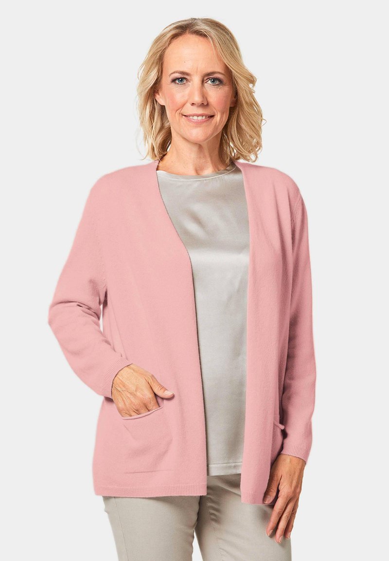 Helles rosa Cardigan mit lockerer Passform, offener Vorderseite und langen Ärmeln. Verfügt über Seitentaschen und gerippte Bündchen, über einem hellgrauen Oberteil getragen.