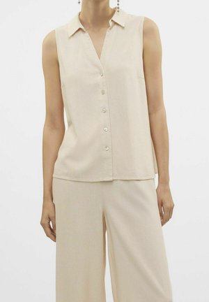 Blusa sin mangas beige con botones y cuello, combinada con pantalones beige sueltos en una persona sobre un fondo liso.