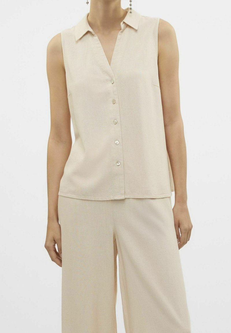 Blouse beige sans manches à boutons avec col, associée à un pantalon beige ample portée par une personne devant un fond uni.