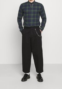 Camisa de cuadros verde y azul marino de mangas largas, combinada con pantalones negros de cintura alta con un detalle de cadena y zapatos negros.