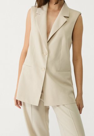 Kvinde iført en ærmeløs beige blazer med knapper og matchende beige bukser mod en ensfarvet lys baggrund.