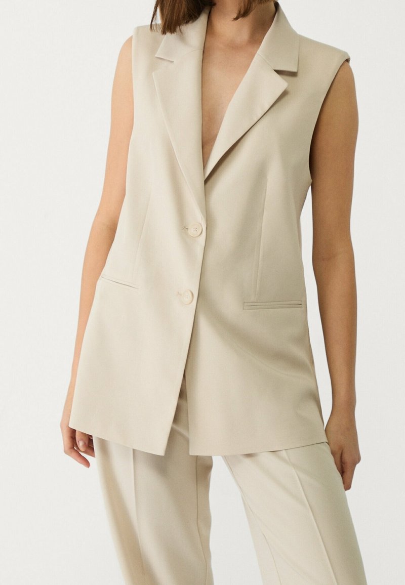 Femme portant un blazer beige sans manches avec des boutons et un pantalon beige assorti sur un fond clair uni.
