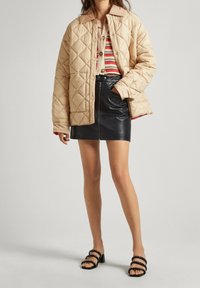 Chaqueta acolchada beige con cuello marrón, llevada sobre una camiseta de rayas rojas y crema, combinada con una minifalda de cuero negra y tacones negros con tiras.