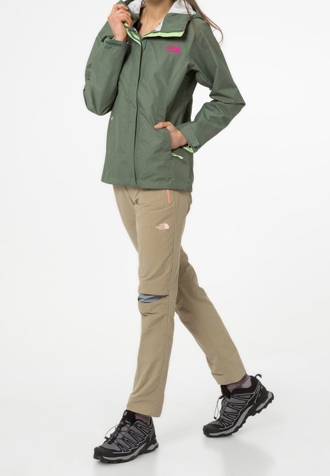 Personne portant une veste verte, un pantalon beige avec des aérations aux genoux et des chaussures de randonnée noires, marchant devant un fond blanc uni.