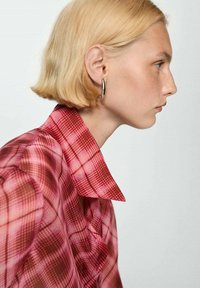 Camicia a quadri rossi con un grande colletto e una morbida texture del tessuto. Il modello ha capelli biondi corti e indossa un orecchino a cerchio di argento.