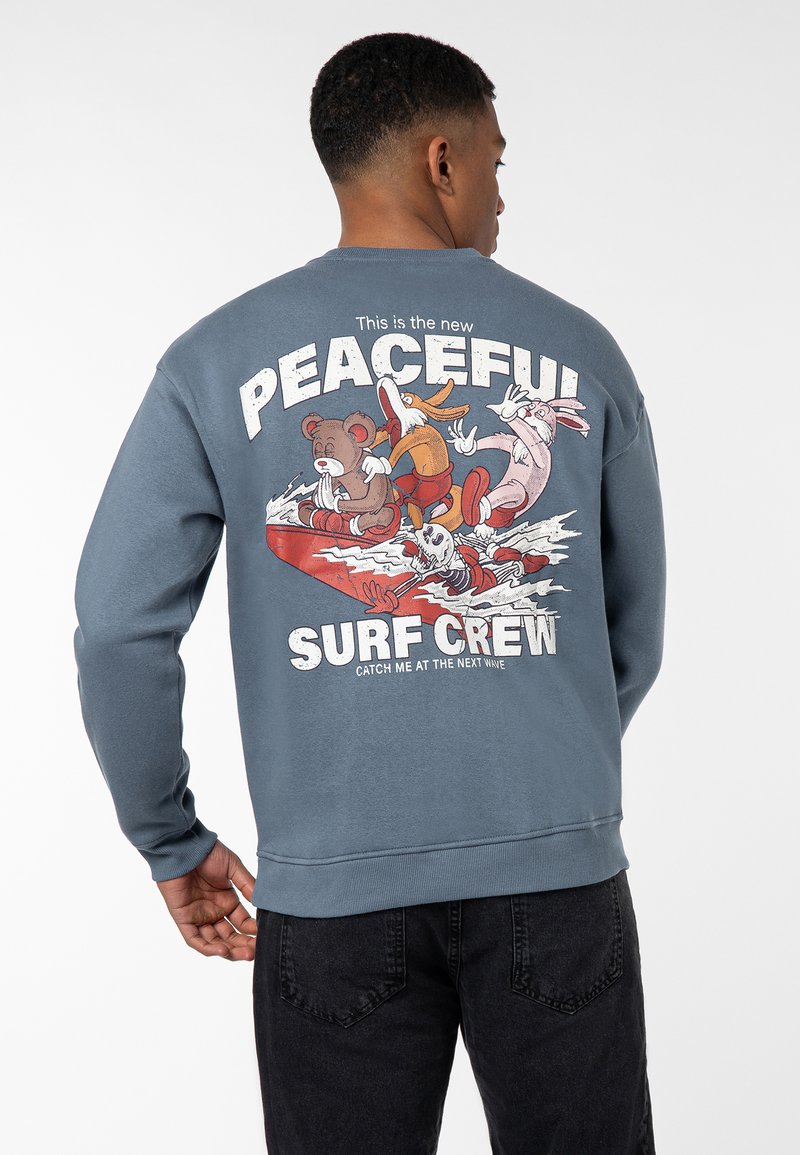 Blue sweatshirt featuring a graphic print of a cartoon bear and friends on a surfboard, with text "This is the new PEACEFUL SURF CREW."
